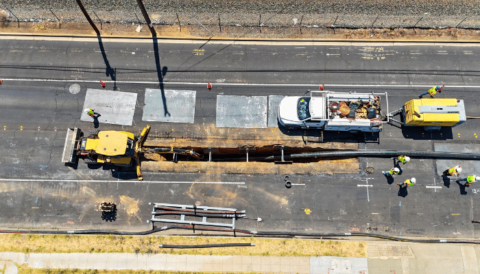 Aerial view of a paved road under construction with a yellow excavator, white truck, and workers near equipment and barriers